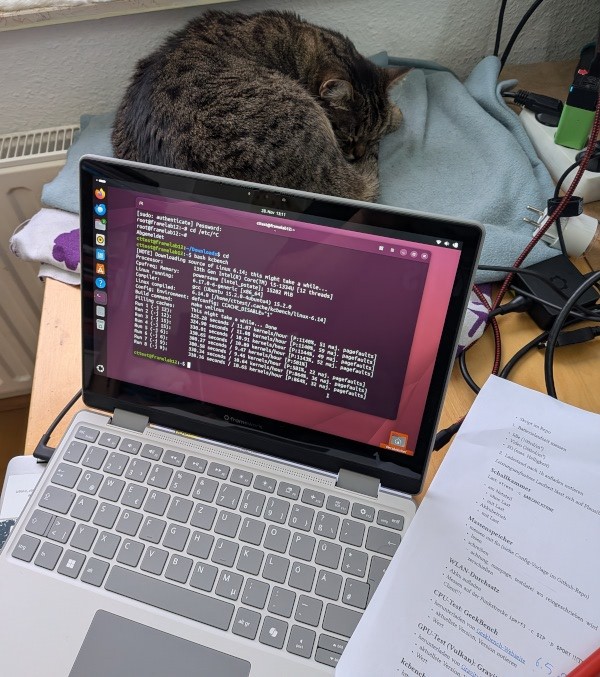 Framework 12 im Laptop-Modus, kcbench lief gerade im Terminal, Test-Arbeitsplan liegt auf Papier daneben, während die Katze ungerührt weiter schläft.