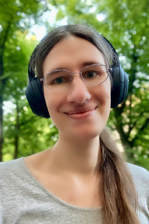 Selfie/Profilfoto von Sophie, junge Frau mit dunkelblonden Haaren lächelt in die Kamera, Brille, dicke Overear-Kopfhörer, Sonnenschein. Im Hintergrund Bäume