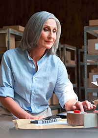Eine Dame mit grauen Haaren arbeitet in einer Lagerhalle an einem Laptop. Foto: Ground Picture/Shutterstock.com.