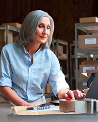 Eine Dame mit grauen Haaren arbeitet in einer Lagerhalle an einem Laptop. Foto: Ground Picture/Shutterstock.com.
