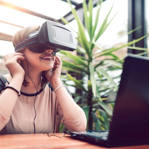 Eine Frau, die eine VR-Brille aufgesetzt hat und damit vor einem Laptop sitzt