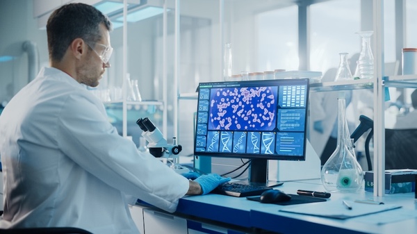A man with protective glasses and gloves in a lab coat sits in a laboratory in front of a computer.