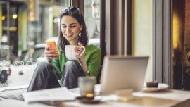 A young woman sitting in a café, holding a cup of coffee and looking at her smartphone, with a laptop on the table