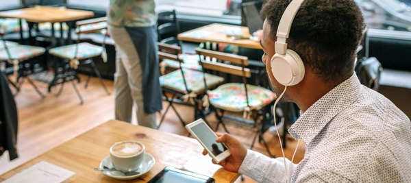 A man wearing headphones sits in a café, looking at his smartphone beside a cup of coffee.