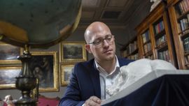A man reading a book in a library, with bookshelves and oil paintings in the background