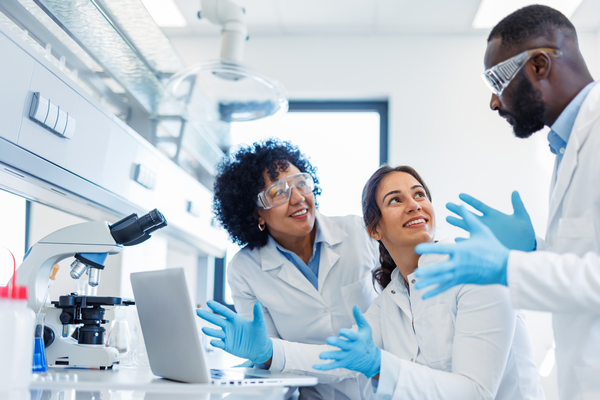 Three people talking in a laboratory. They are wearing lab coats, medical gloves and safety glasses.
