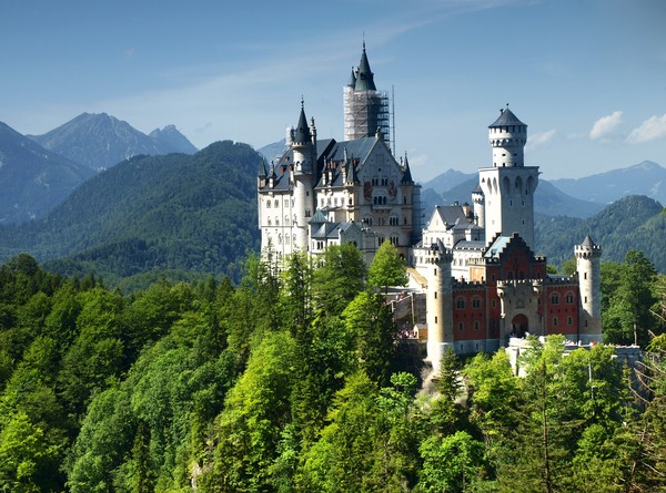 A castle nestled in the woods on a mountain, with an alpine panorama in the background.