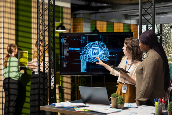 Two women stand in an office and look at a screen showing a digital brain visualisation with the word AI