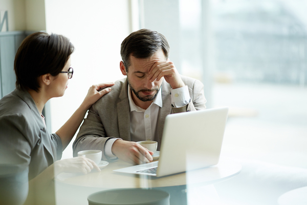 Concerned man holding his forehead at a laptop while a colleague comforts him