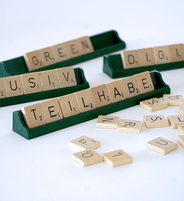 Auf einem weißen Tisch liegen einige Buchstabensteine und vier Ablageleisten eines Wortlegespieles. Auf den Ablageleisten sind die Worte "Inklusiv", "Teilhabe", "Green" und "Digital" zu lesen.