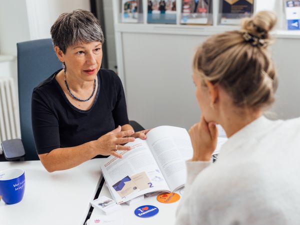 Beratungssituation an einem Schreibtisch; eine ältere Frau hat ein Prospekt aufgeschlagen und zeigt einer gegenüber sitzenden, jüngeren Frau etwas darin.