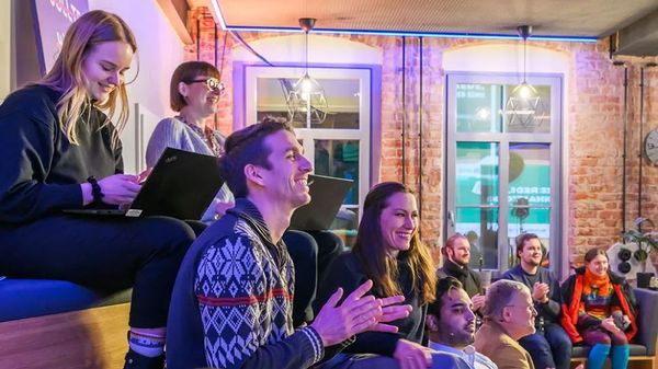 Eine Gruppe junger Erwachsener sitzt in einem modern eingerichteten Raum mit Backsteinwänden und hört aufmerksam zu. Einige lächeln und applaudieren, andere machen sich Notizen auf Laptops. Die Atmosphäre wirkt offen, engagiert und gemeinschaftlich, wie bei einer Präsentation oder einem Austauschformat.