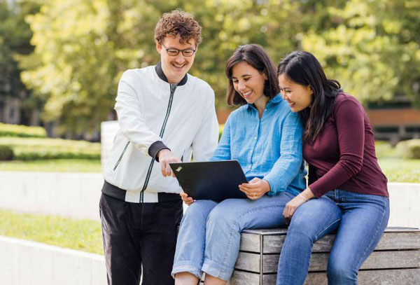 Drei junge Erwachsene sitzen im Freien auf einer Holzbank und schauen gemeinsam lächelnd auf einen Laptop. Sie befinden sich in einer grünen Umgebung, die an einen Campus oder Park erinnert, und wirken entspannt und konzentriert.