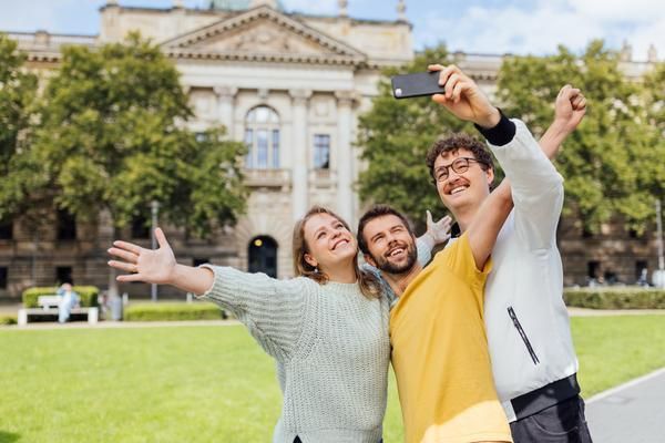 Drei junge Erwachsene stehen auf einer Wiese vor einem historischen Gebäude und machen ein fröhliches Selfie. Sie lächeln in die Kamera, haben die Arme ausgebreitet und wirken ausgelassen an einem sonnigen Tag.