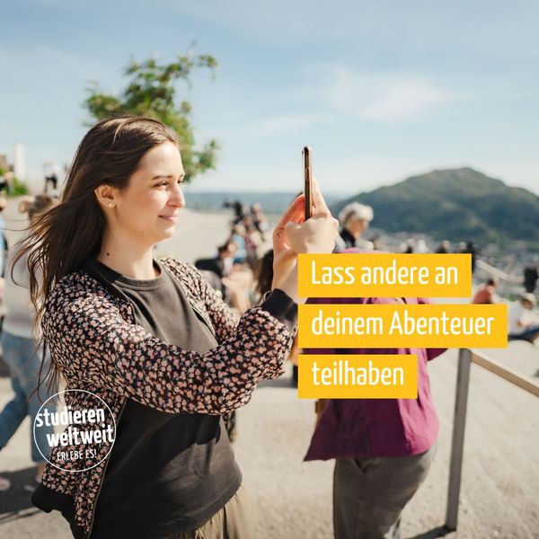 Junge Frau fotografiert mit einem Smartphone von einem Aussichtspunkt. Sie blickt auf den Bildschirm, im Hintergrund eine unscharfe Naturkulisse.