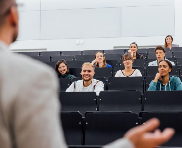 Zu sehen ist eine Hörsaal-Szene: ein Mann steht auf dem Podium und spricht, während ein kleines Publikum interessiert zuhört.
