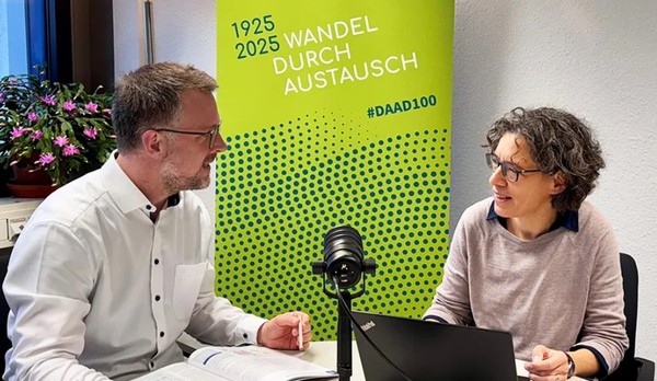 Pressesprecher und Direktorin Kommunikation des DAAD sitzen sich an einem Tisch gegenüber und sprechen in ein Mikro.