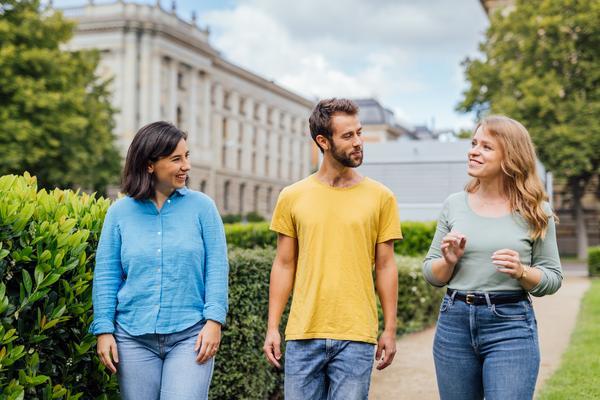 Drei Menschen spazieren über ein Hochschulgelände und tauschen sich lächelnd miteinander aus.