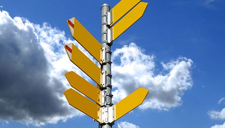Ein Schild-Pfosten mit mehreren, gelben Wegweiser-Pfeilen in unterschiedliche Richtungen steht vor blauem Himmel mit einzelnen Wolken.