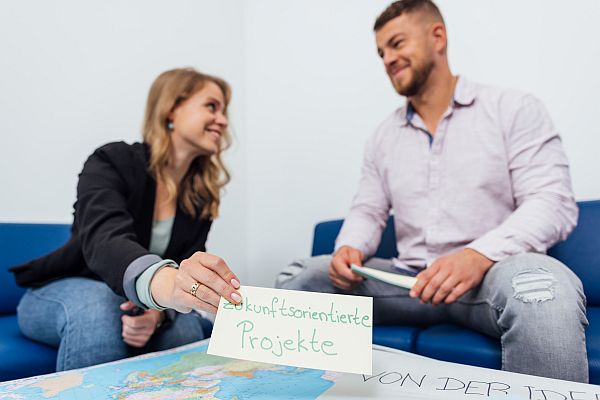Zwei Personen sitzen sich auf einem Sofa gegenüber und lächeln sich an. Die Frau hält eine Karte mit der Aufschrift „zukunftsorientierte Projekte“ über eine Weltkarte auf einem Tisch, während der Mann ein Notizbuch in der Hand hält.