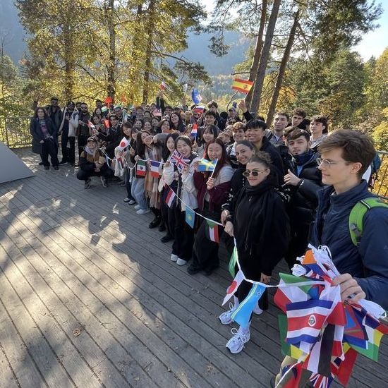 Eine große Gruppe Studierende, die sich ehrenamtlich für Europa engagieren, steht auf einer Aussichtsplattform vor Bäumen und Bergen im Hintergrund. Sie halten gemeinsam eine Girlande mit vielen unterschiedlichen Flaggen, die sie miteinander verbindet.