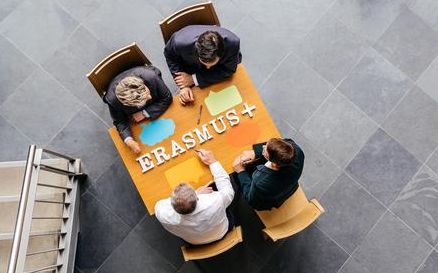 Vogelperspektive auf vier Personen, die an einem Tisch sitzen. Auf der Tischplatte liegt der Schriftzug „ERASMUS+“ aus Holzbuchstaben. Die Personen reichen sich über den Tisch die Hand und symbolisieren internationale Zusammenarbeit und Partnerschaft im Rahmen von Erasmus+.