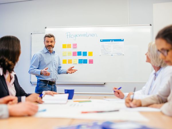 Ein Mann steht vor einem Whiteboard mit Text und bunten Haftnotizen und erklärt etwas. Vor ihm sitzen an Tischen einige Zuhörer.