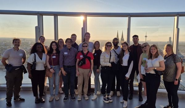 Gruppenfoto der ehrenamtlichen Helfer beim Netzwerktreffen in Köln. Sie stehen vor einer Glasfront auf einer Aussichtsplattform, im Hintergrund sieht man die Stadt Köln, den Rhein und den Kölner Dom.