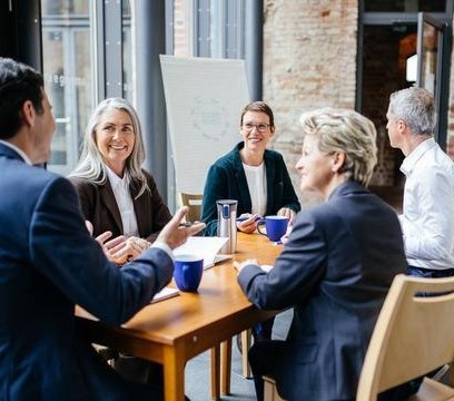 Fünf Personen sitzen gemeinsam an einem Besprechungstisch und tauschen sich in einer modernen, hellen Arbeitsumgebung aus. Im Hintergrund sind ein Flipchart und große Fenster zu sehen.
