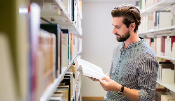 Ein junger Mann liest in einem Buch zwischen zwei Bibliotheksregalen.