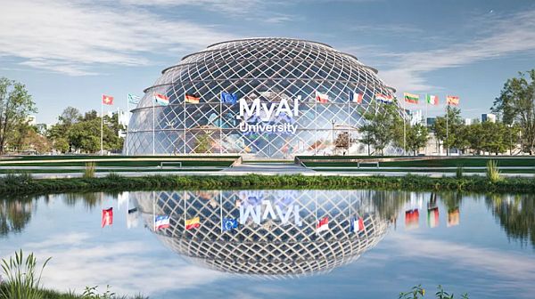 Modernes, gläsernes Kuppelgebäude der „MVA University“ mit geometrischer Gitterfassade, davor ein Wasserbecken mit Spiegelung. Vor dem Gebäude wehen mehrere europäische Nationalflaggen und die EU-Flagge, umgeben von Grünflächen und Bäumen unter blauem Himmel.
