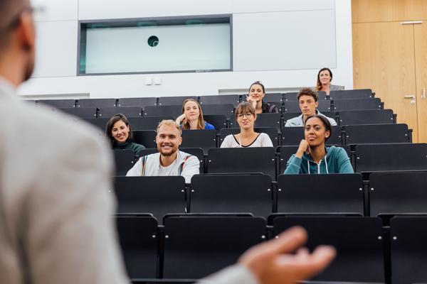 Zu sehen ist eine Hörsaal-Szene: ein Mann steht auf dem Podium und spricht, während ein kleines Publikum interessiert zuhört.