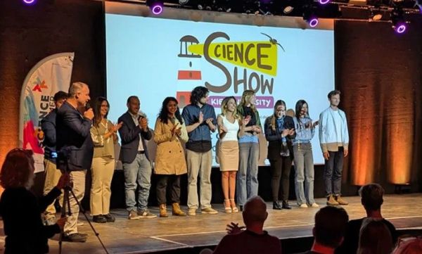 Eine Gruppe junger Erwachsener steht auf einer Bühne und applaudiert gemeinsam mit dem Publikum. Hinter der Bühne ist auf einer Leinweinwand ein Leuchtturm und der Schriftzug "Science Show" zu erkennen.