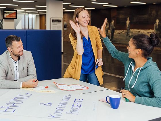 Drei fröhliche Personen sitzen und stehen an einem Tisch, auf dem Poster und Werbematerialien liegen - zwei von ihnen geben sich enthusiastisch einen High-Five.
