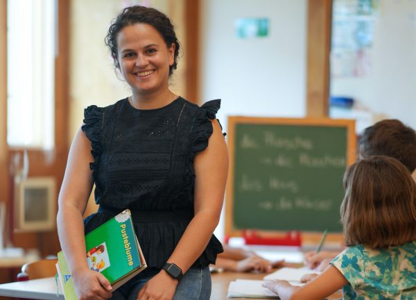 Junge Frau in einem Klassenzimmer, im Hintergund sind Schüler und eine Tafel zu erkennen.