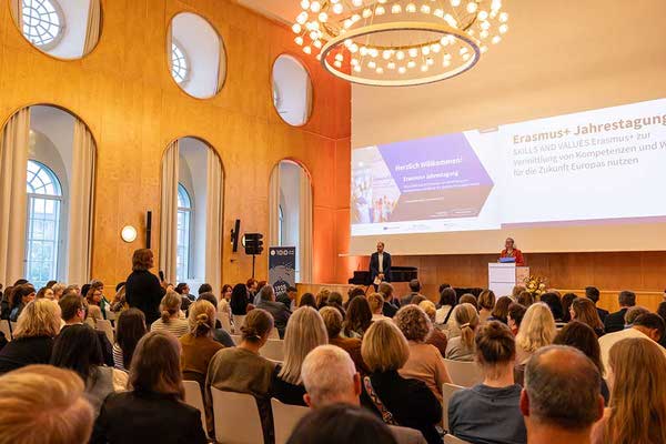 Blick in den Saal der Erasmus+ Jahrestagung mit Publikum, Redner*innen auf der Bühne und einer stehenden Teilnehmerin, die gerade ein Mikrofon hält und eine Frage stellt.