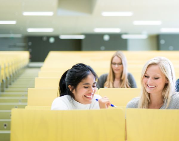 Zwei junge Frauen sitzen in einem hellen Hörsaal - sie sind sich zugewandt und lächeln.