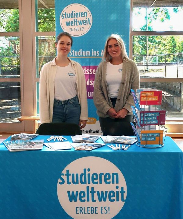 Zwei junge Frauen stehen hinter einem Infostand von "studieren weltweit - ERLEBE ES!". Sie sind bereit für Besucher und deren Fragen.