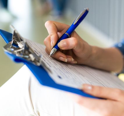 Nahaufnahme einer Person, die mit einem blauen Kugelschreiber ein Formular auf einem Klemmbrett ausfüllt. Das Bild symbolisiert Antragstellung, Dokumentation oder administrative Verfahren.