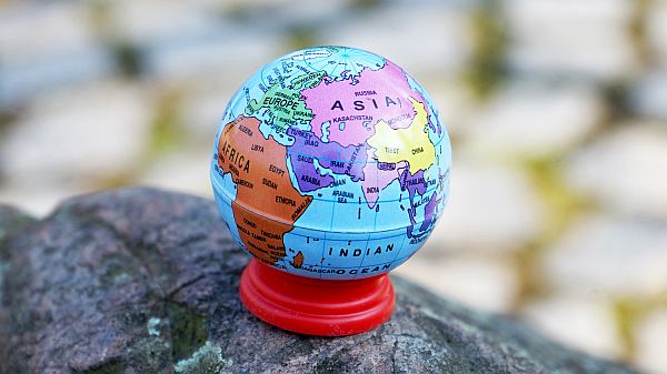 Kleiner bunter Globus mit rotem Sockel steht auf einem Stein. Sichtbar sind Europa, Afrika und Asien mit Beschriftungen der Länder- und Kontinente; der Hintergrund ist unscharf und wirkt natürlich.