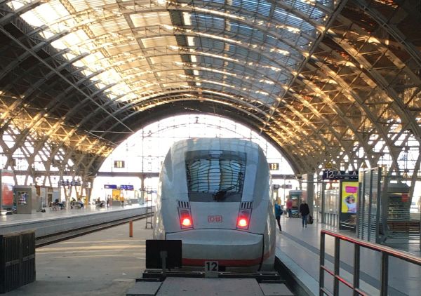 Eine Frau, von Hinten fotografiert, steht an einem S-Bahnsteig. Sie ist in Schwarz gekleidet und hält einen Jutebeutel in ihren Händen. Vor ihr ist eine gelbe Bahn in Bewegung und verschwommen zu erkennen.