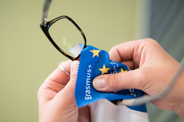 Eine Hand reinigt eine Brille mit einem blauen Erasmus+-Brillenputztuch mit EU-Sternen.