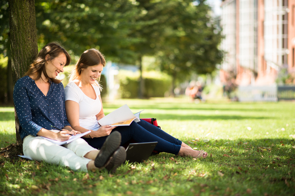Zwei junge Frauen sitzen angelehnt unter einem Baum auf einer Wiese. Die Sonner scheint, sie blicken lächelnd in Bücher und Unterlagen.