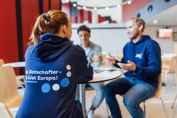 Drei junge Menschen sitzen an einem runden Tisch und sprechen angeregt miteinander. Die tragen Hoodies mit der Aufschrift "Sei Botschafter - Du bist Europa!".