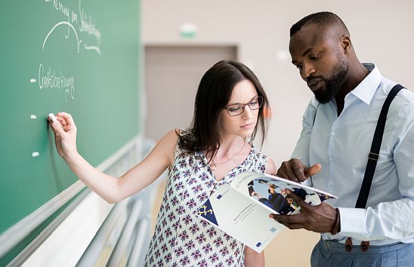 Eine Frau und ein Mann stehen in einem Klassenraum vor einer Tafel. Die Frau zeigt auf handschriftliche Notizen an der Tafel, während der Mann ein aufgeschlagenes Heft hält, in das beide konzentriert schauen. Die Situation wirkt wie eine gemeinsame Besprechung oder Lernsituation.