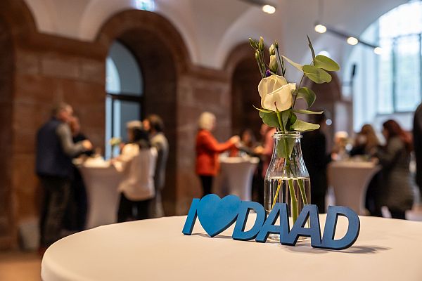 Zu sehen ist unscharf im Hintergrund ein Foyer mit hohen Decken und großen Fenstern. Im Vordergrund stehen eine Blumenvase und ein DAAD-Schriftzug auf einem Stehtisch, während sich im Hintergrund mehrere Personen an weiteren Stehtischen unterhalten.