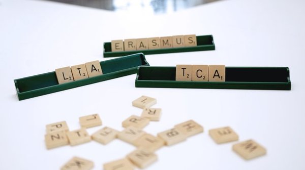 Scrabble-Steine liegen auf einem Tisch; in einer Ablageleiste ist das Wort „ERASMUS“ zu lesen, auf dem Tisch „LTA“ und „TCA“ gelegt. Weitere Buchstabensteine liegen ungeordnet davor.