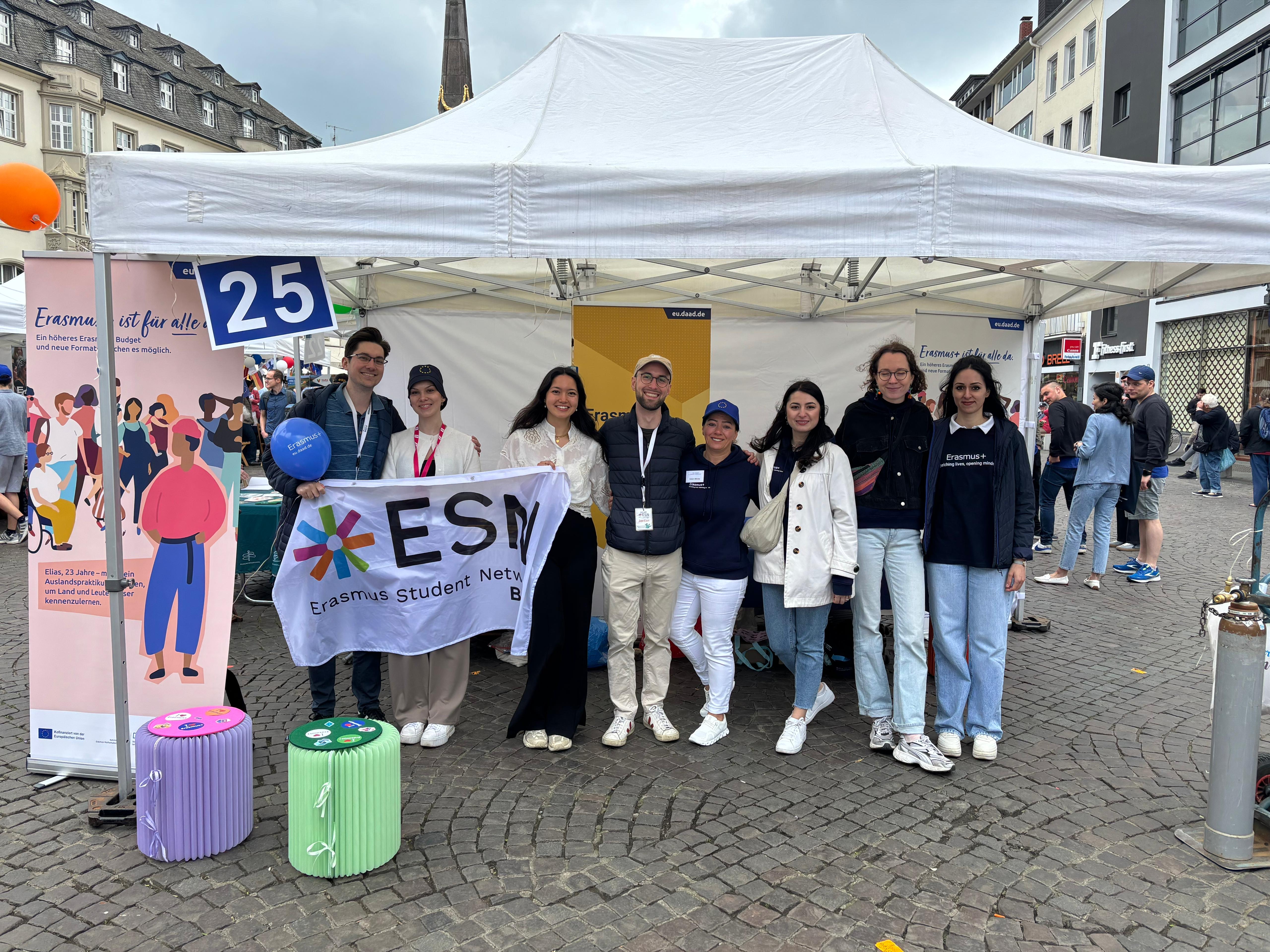 Zu sehen sind DAAD-Kolleginnen und ESN-Mitglieder am gemeinsamen Stand auf dem Bonner Marktplatz beim Europatag Bonn. 3 Personen halten eine Fahne hoch.