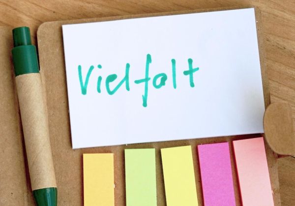 Auf einem Holztisch liegt ein kleines Heftchen mit einem weißen Haftnotiz-Block, bunten Haftmarkern und einem Kugelschreiber. Auf der Haftnotiz ist das Wort "Vielfalt" zu lesen.