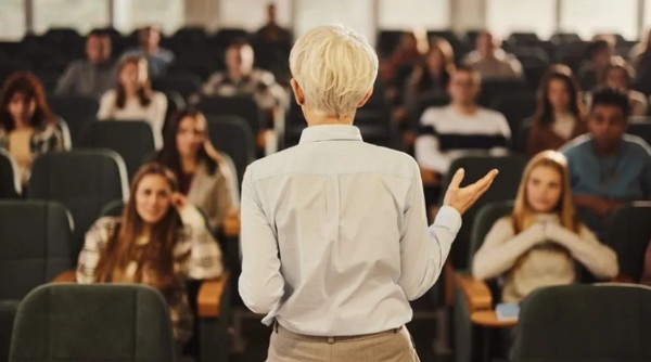 Zu sehen ist eine Hörsaal-Szene: eine Frau steht auf dem Podium und spricht, während eine Gruppe Studierende interessiert zuhört.